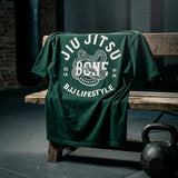 Dark green BJJ Lifestyle oversize t-shirt with snake Breaking Bone graphic on a bench in a martial arts gym.