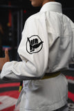 Close-up of a Kids White BJJ Gi jacket featuring a black shaka hand patch, pearl weave fabric, and reinforced stitching.